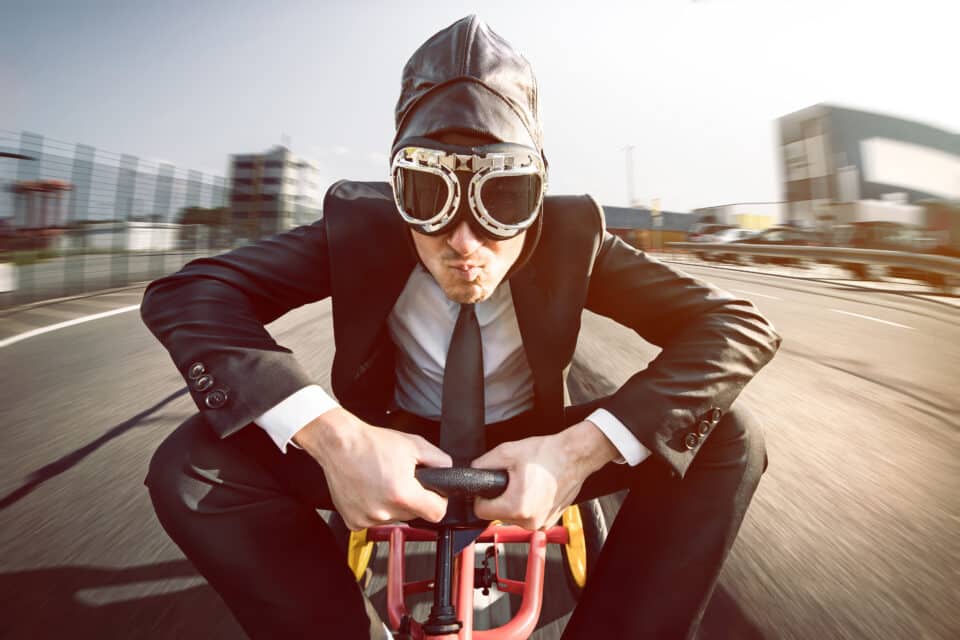 Mann mit Anzug und Fliegerbrille sitzt auf Tretauto