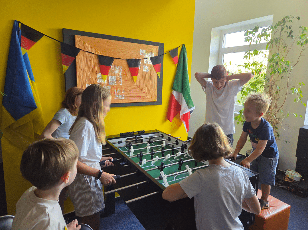 Das Bild zeigt Kinder, die vergnügt Tischfussball spielen, vor einer Wand mit verschiedenen Flaggen, passend geschmückt zur WM
