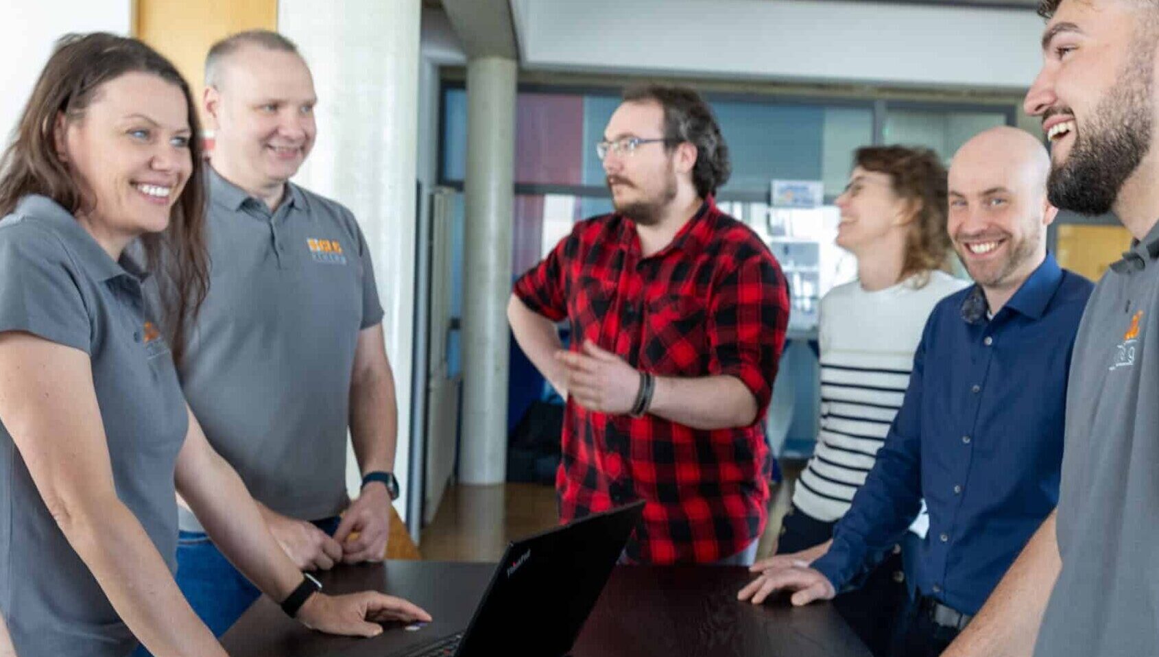 6 employees of CLC xinteg GmbH stand happily together at a table and work with a laptop